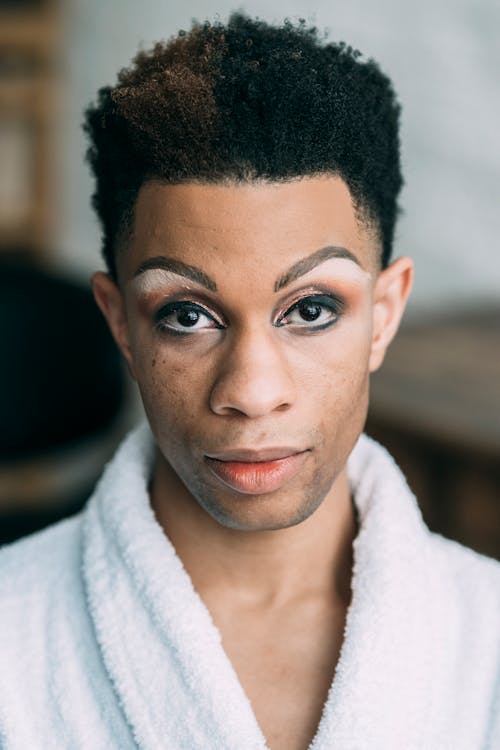 Free Serious feminine African American male with bright eyeshadow wearing bathrobe looking at camera while standing in room on blurred background Stock Photo