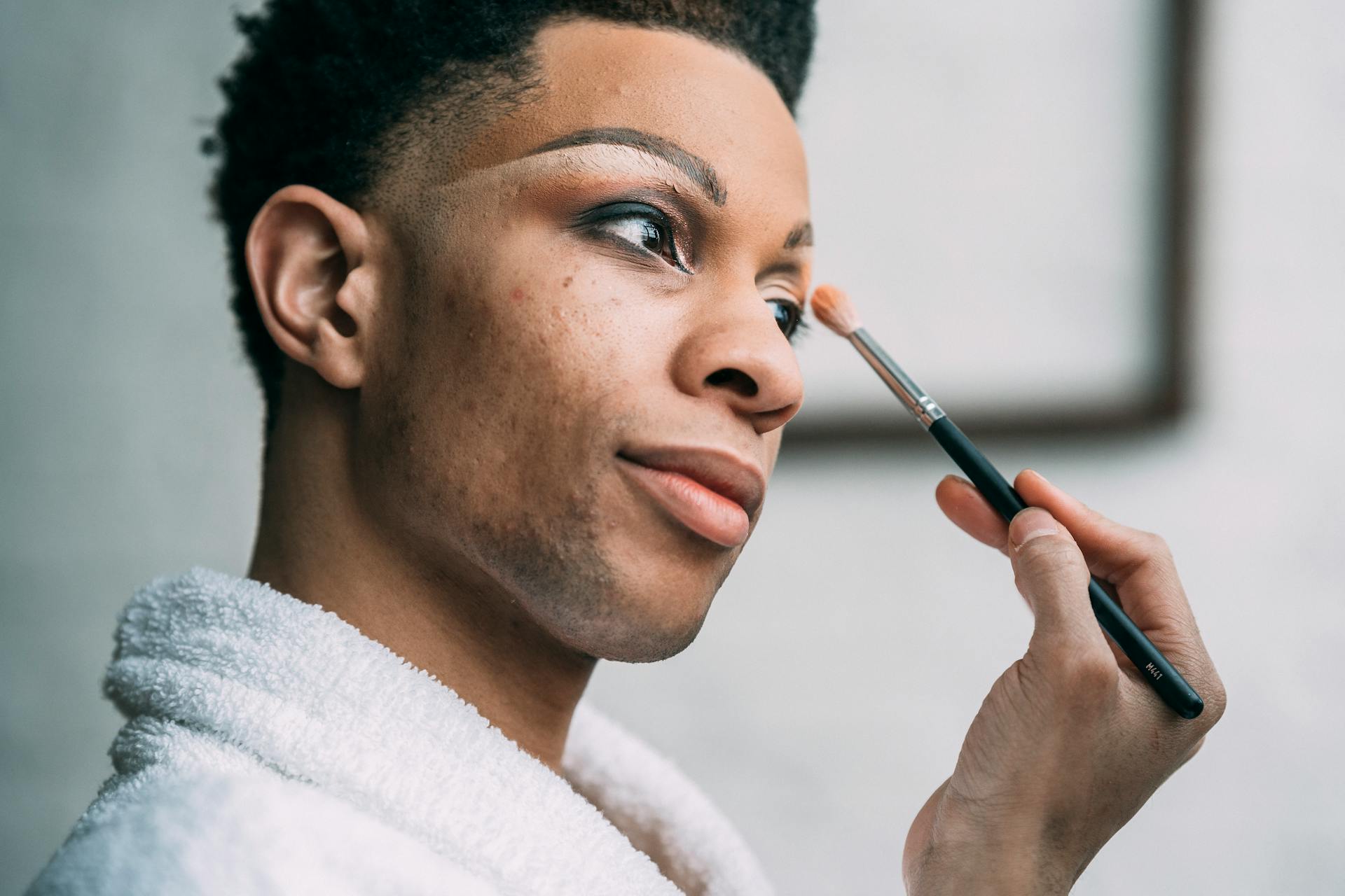 Side view of queer African American male in white bathrobe applying eyeshadow with cosmetic brush in light room at home
