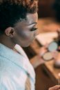 Side view of eccentric African American male with bright makeup sitting at table with various cosmetics in modern dressing room