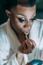 Feminine African American male with dark hair in white bathrobe with eyelashes applying lip pencil on lips in dressing room