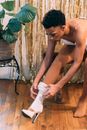 African American young male in drag queen costume sitting on wooden bench while dressing half hose on heels in light dressing room near potted plant