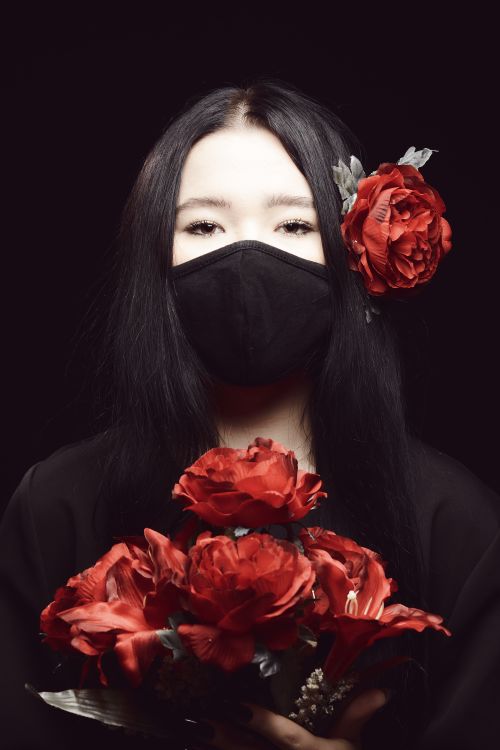 Free Elegant portrait of a woman wearing a face mask, holding red roses, set against a black background. Stock Photo