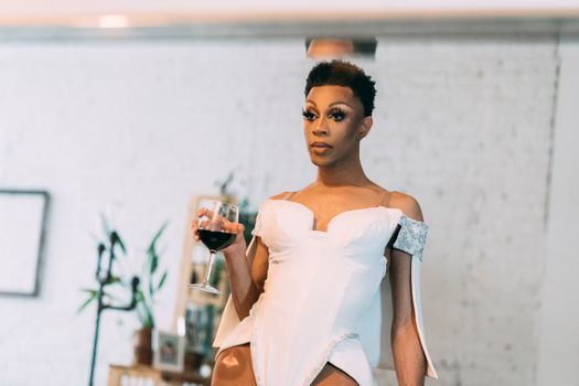 Calm African American drag queen in elegant outfit holding glass of red wine looking at mirror in light room