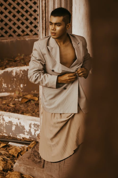 Stylish ethnic male model wearing formal jacket and maxi skirt standing in desolated building patio and looking away