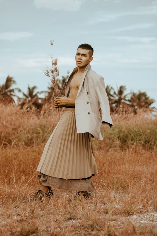 Free Full body young ethnic male model wearing formal jacket on bare chest and maxi skirt while standing on grassy valley and looking at camera Stock Photo