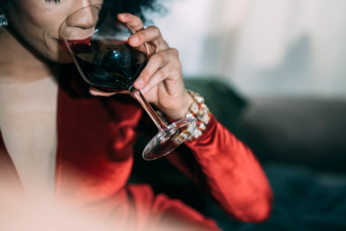 Crop unrecognizable female in bright red dress with jewelry drinking red wine on blurred background