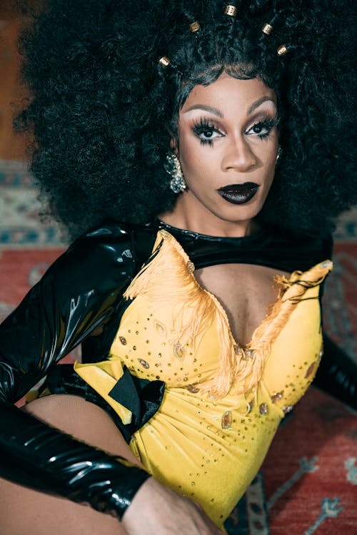 Self assured young black male drag queen in wig and bright makeup wearing yellow and black costume lying on carpet