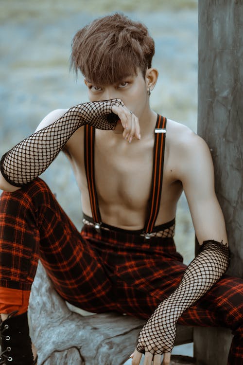 Free Unemotional extravagant male with bare chest wearing checkered trousers and net fingerless mittens touching face and looking at camera while sitting on tree trunk in garden Stock Photo