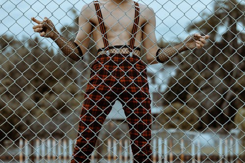 Crop faceless shirtless man in checkered trousers behind net fence