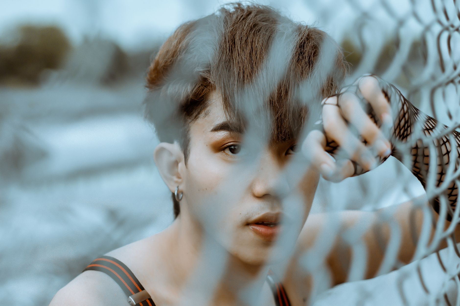 Unemotional shirtless ethnic male with makeup wearing fingerless gloves looking at camera through net fence
