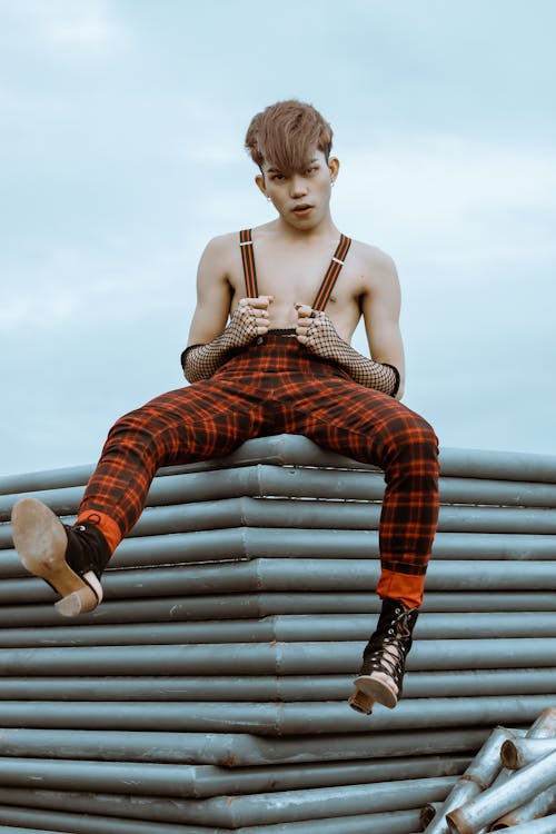 Free Full body confident extraordinary ethnic male with bare chest wearing checkered trousers and high heeled shoes sitting on metal construction against blue sky Stock Photo