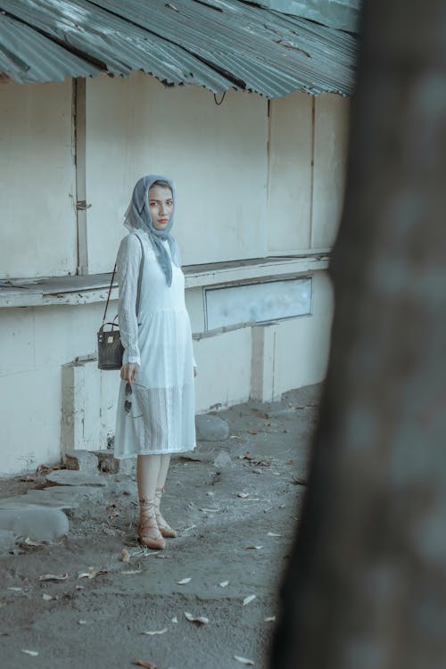 Full length unemotional ethnic female in white dress and headscarf standing near shabby building on street and looking at camera
