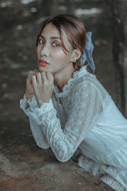 Sensitive young ethnic female wearing white lace dress leaning on hands and looking at camera while sitting at weathered wooden table in garden