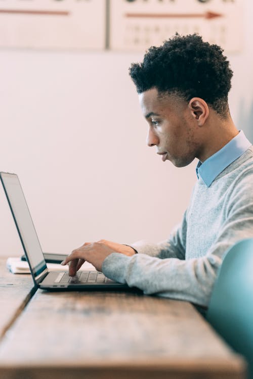 Free Side view of concentrated ethnic bearded male with short curly hair sitting at wooden desk and looking at screen of laptop while typing Stock Photo