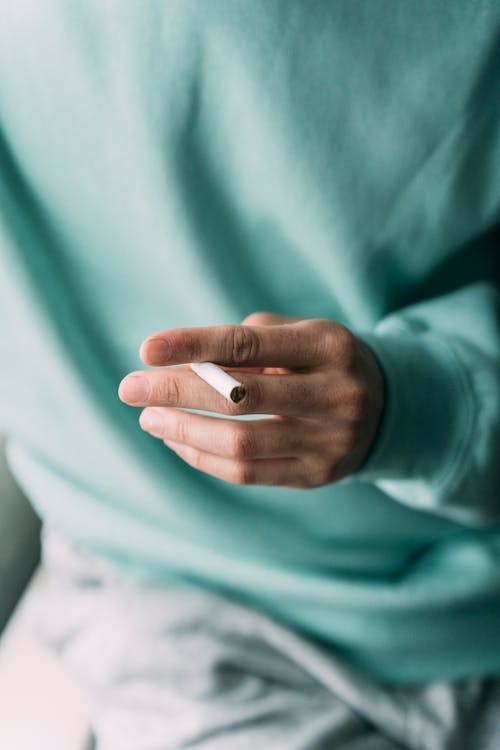 Crop view of ethnic anonymous person wearing light sweater and holding cig with fingers in bright daylight
