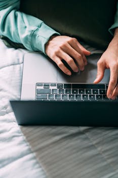 From above crop unrecognizable person using laptop while lying on comfortable bed with blanket in soft daylight