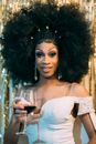 Cheerful black male drag queen with bright makeup in white elegant clothes and curly black wig holding glass of red wine and looking at camera against light golden background