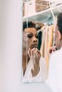 Reflection of black man in white gown standing in front of mirror and doing makeup with lip pencil