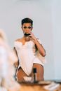Pensive African American drag queen performer with bright makeup and stylish clothes standing near mirror and drinking glass of red wine in light dressing room