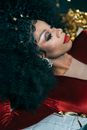 Gorgeous African American drag queen with bright makeup and hairstyle in fashionable clothes lying with closed eyes and leaning on hand in during performance in festive studio