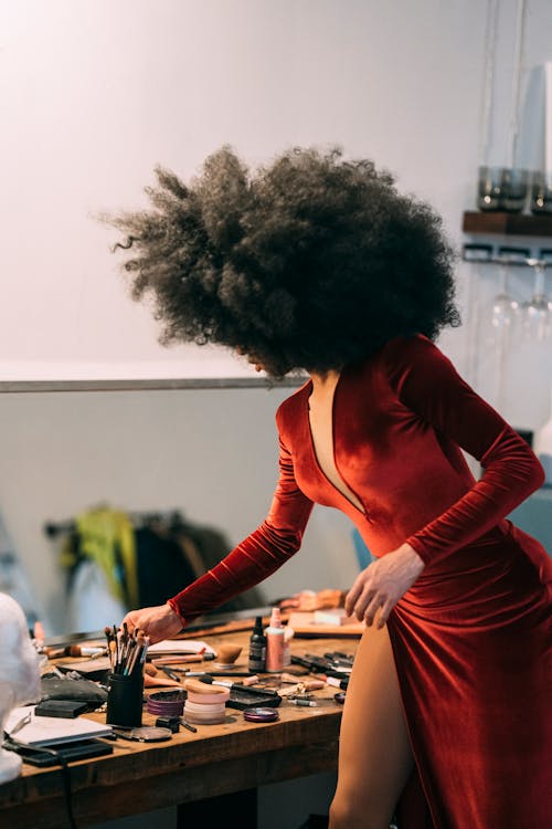 Side view of stylish unrecognizable African American model with black curly hair in long red dress standing near table and taking cosmetic in dressing room