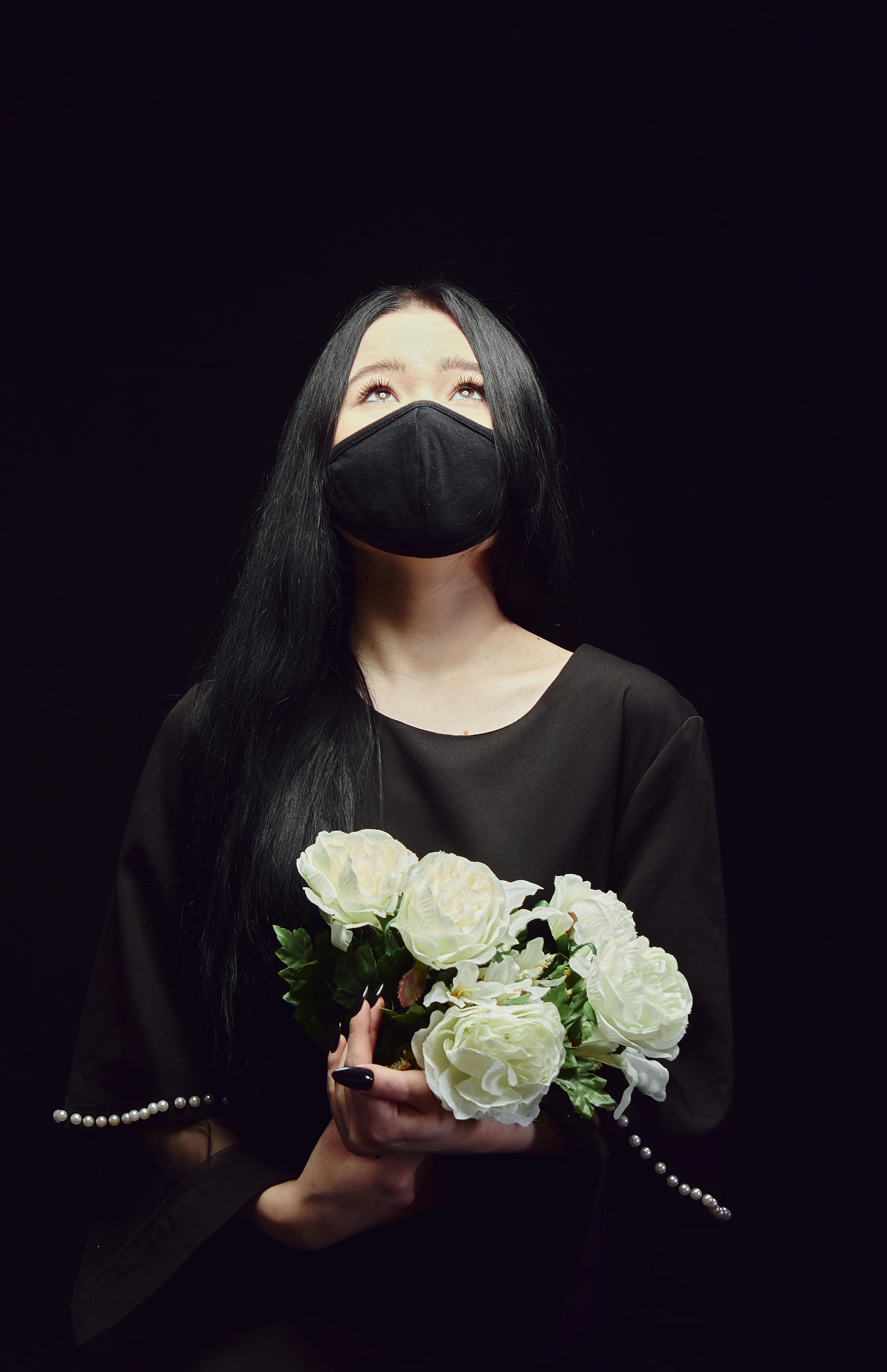 A woman in black attire holds a bouquet of white roses, wearing a mask and looking upwards against a dark background.