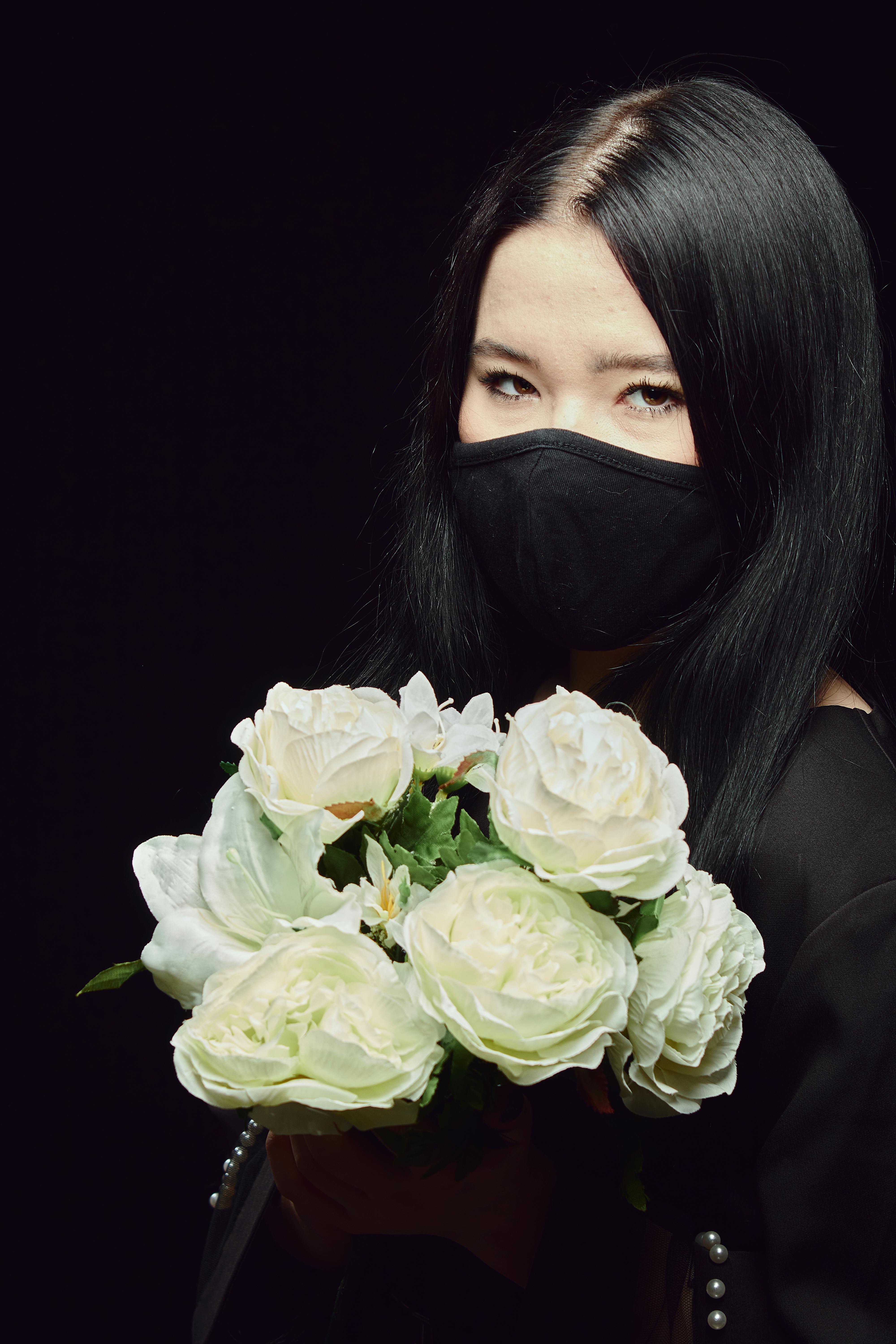 A woman in a black mask holds a bouquet of white roses against a dark background.