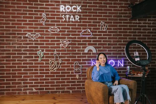 Free Asian woman streaming content with neon signs on a brick wall. Modern influencer setup. Stock Photo