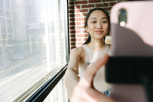 Smiling Asian woman taking a selfie near a large window with brick wall background.