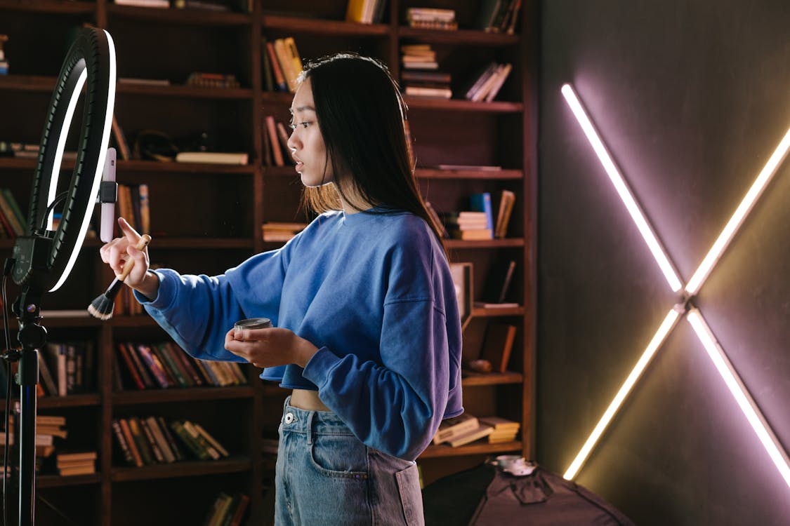 Woman in Blue Long Sleeve Shirt and Blue Denim Jeans Standing Beside Brown Wooden Shelf