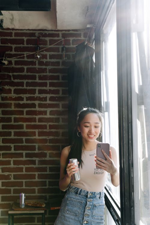 Free Asian woman enjoys a soda while taking a selfie in a brick-walled loft. Stock Photo