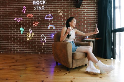 Free Young woman in stylish casual wear taking a selfie in a chic room with neon decor and brick walls. Stock Photo