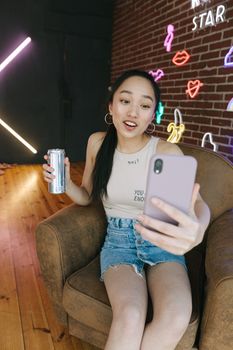 Asian woman taking selfie with smartphone while sitting on a sofa in a neon-lit room with a drink.