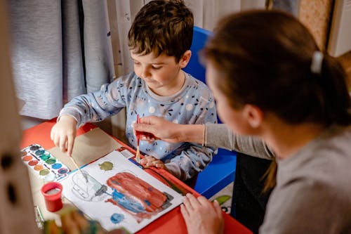 High angle of mum with paintbrush helping son in drawing picture with watercolor paints in kids bedroom
