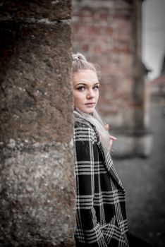 Portrait of a woman in a plaid coat leaning against a brick wall outdoors.