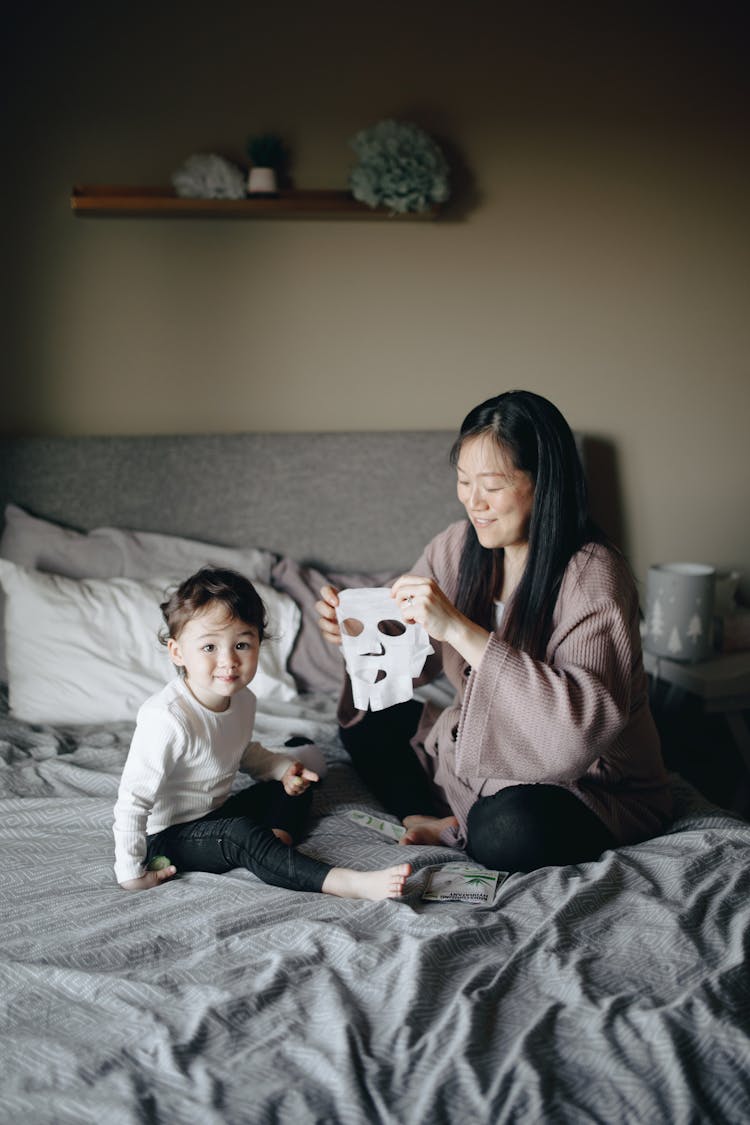 Mother Holding A Mask While Sitting On Bed With Her Child