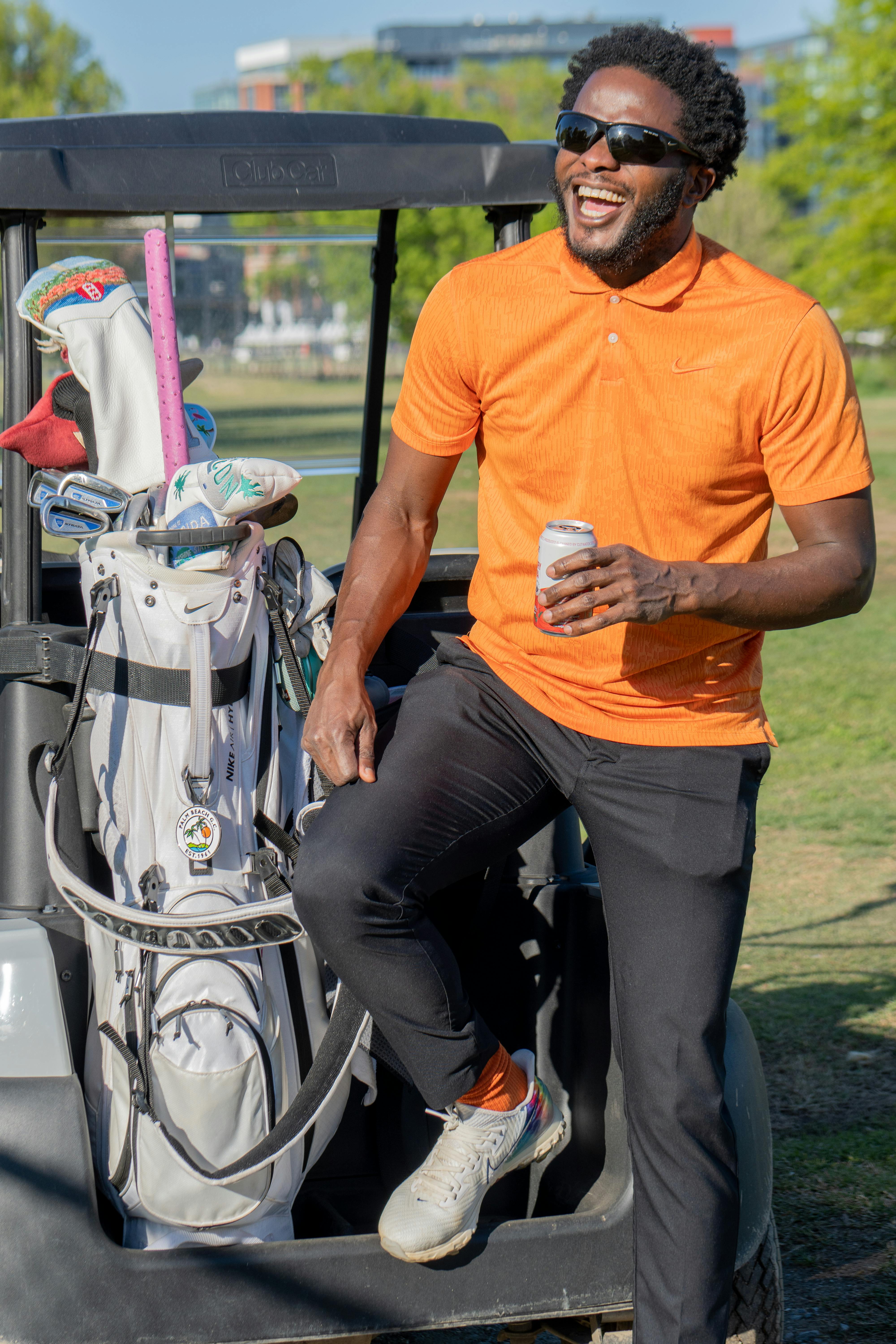 Smiling man in orange shirt holding a drink next to a golf cart outdoors.