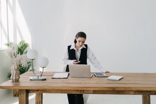 A businesswoman working at a modern wooden desk with a laptop and tablet in a bright office space.
