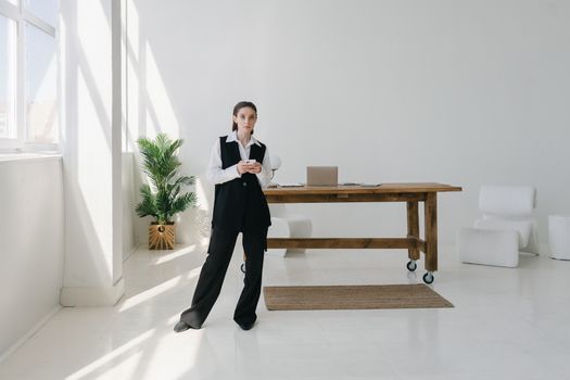 A young woman in a modern office setting uses a smartphone while leaning against a desk.