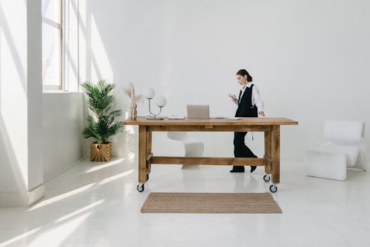 Elegant minimalist office interior with wooden table, potted plant, and woman using smartphone.