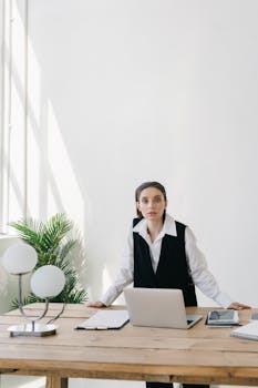 Professional woman working at a desk with laptop, tablet, and lamp in a bright office.