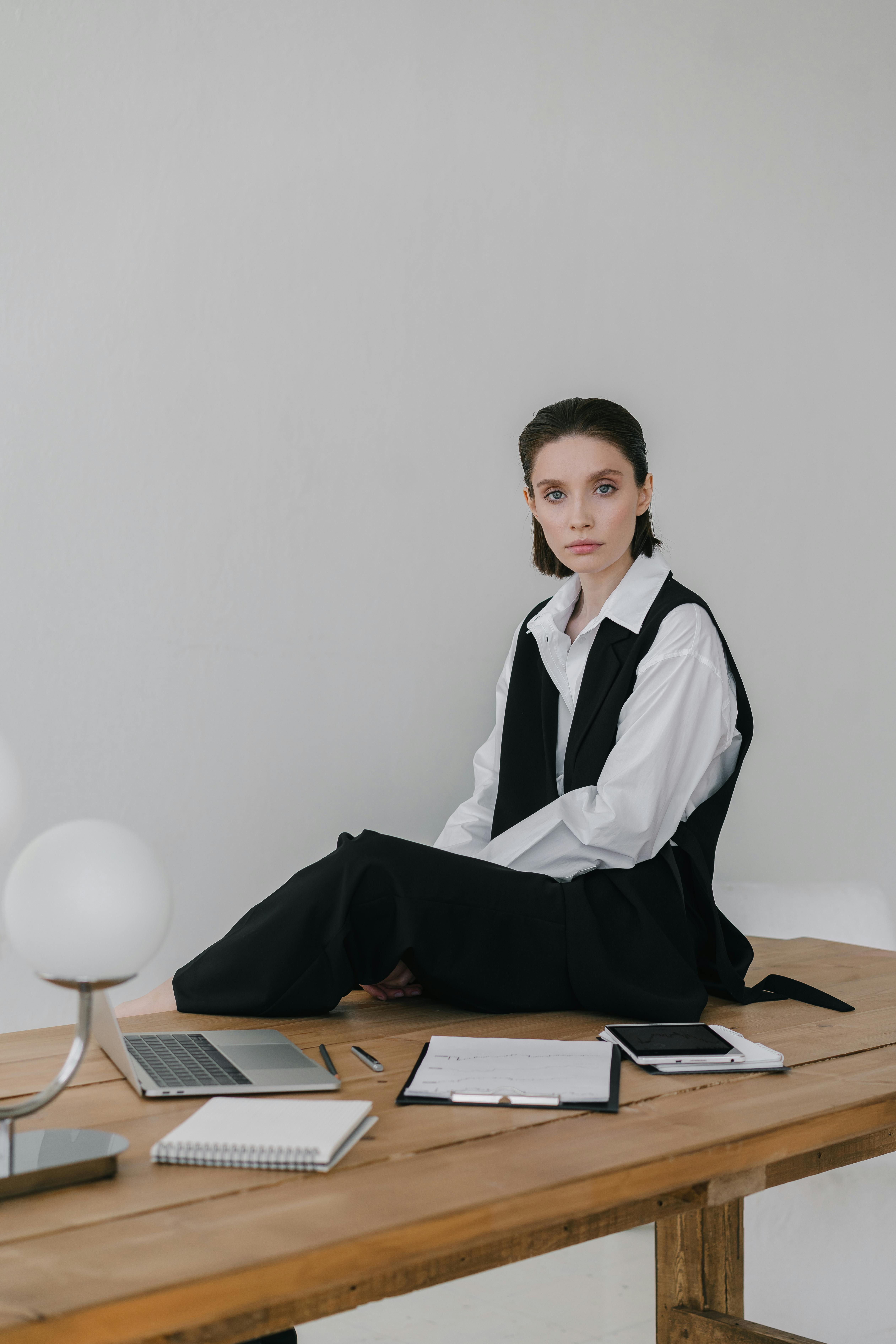 a woman wearing a vest and a white long sleeved shirt sitting on a wooden desk