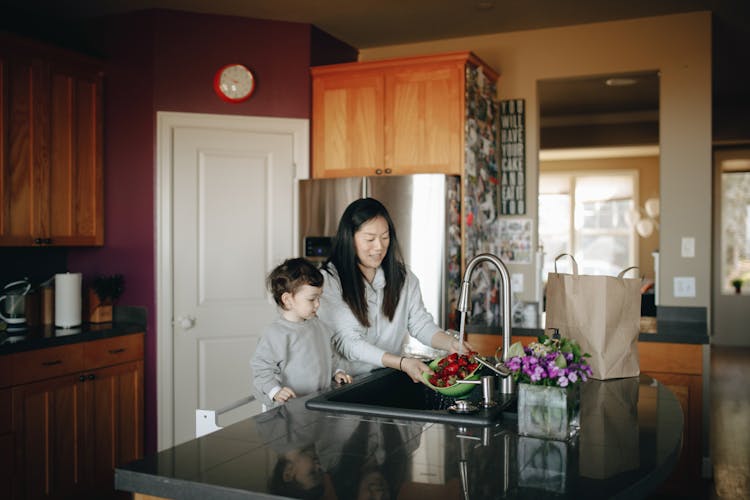 Mother Washing Strawberries Beside Her Child In The Kitchen