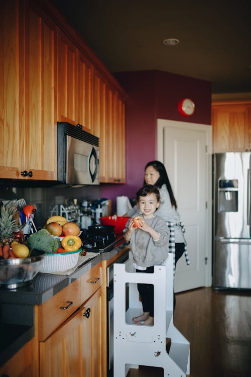 Adorable Child Standing On A Stool In The Kitchen With Her Mother