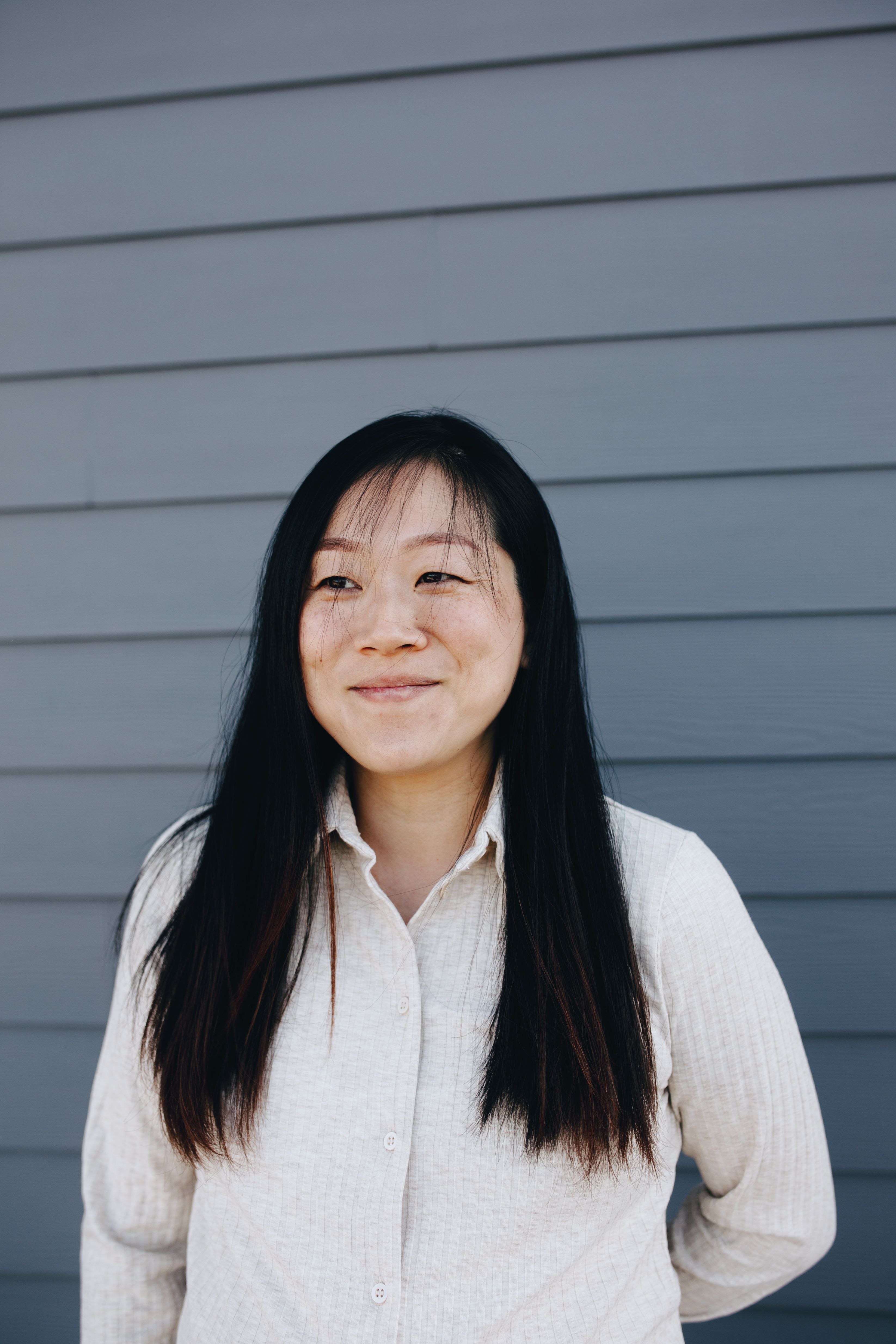A woman with long black hair smiles softly while standing against a gray wall.