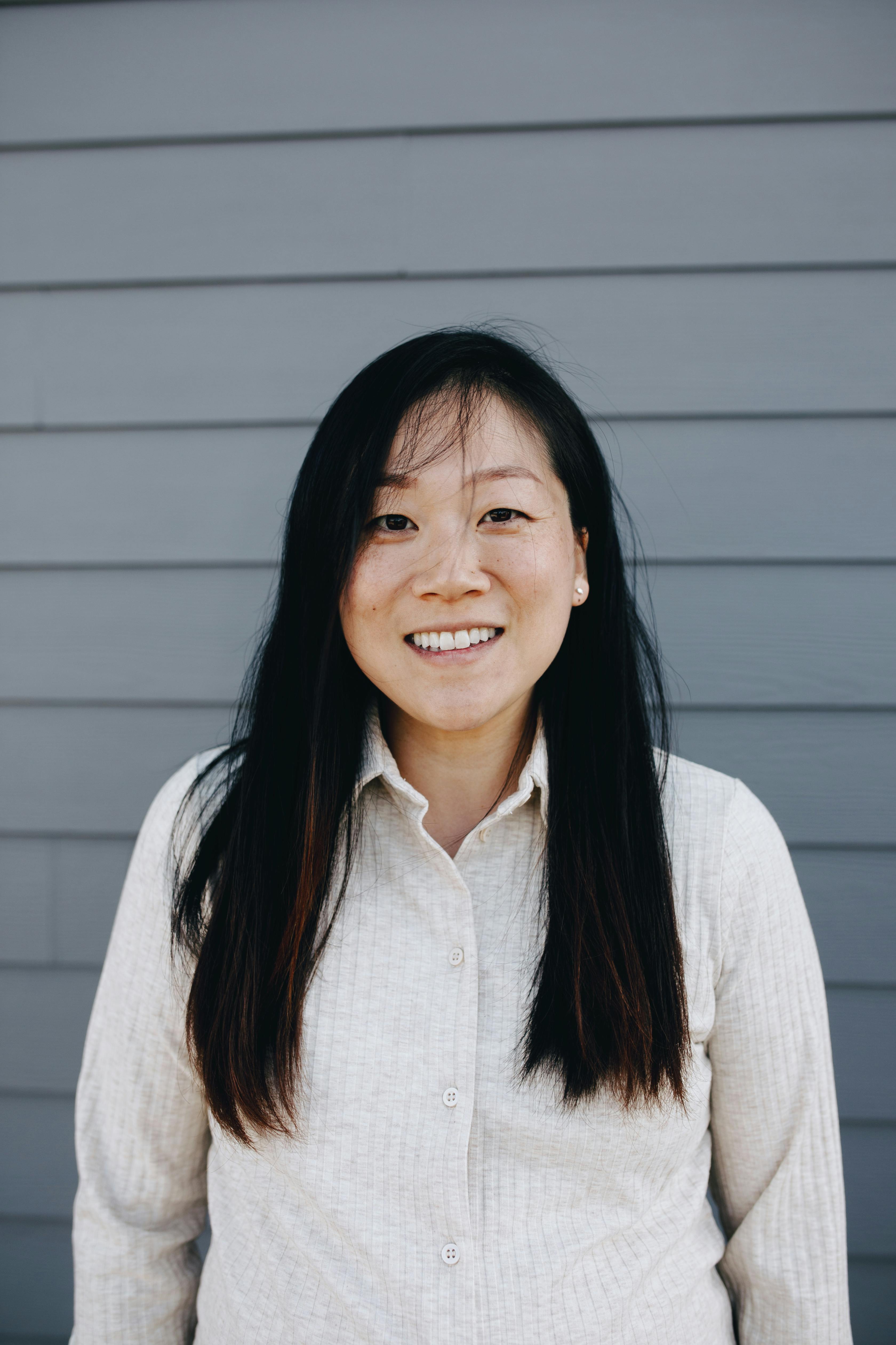 Portrait of a smiling pregnant Asian woman in casual knitwear against a gray wall outdoors.