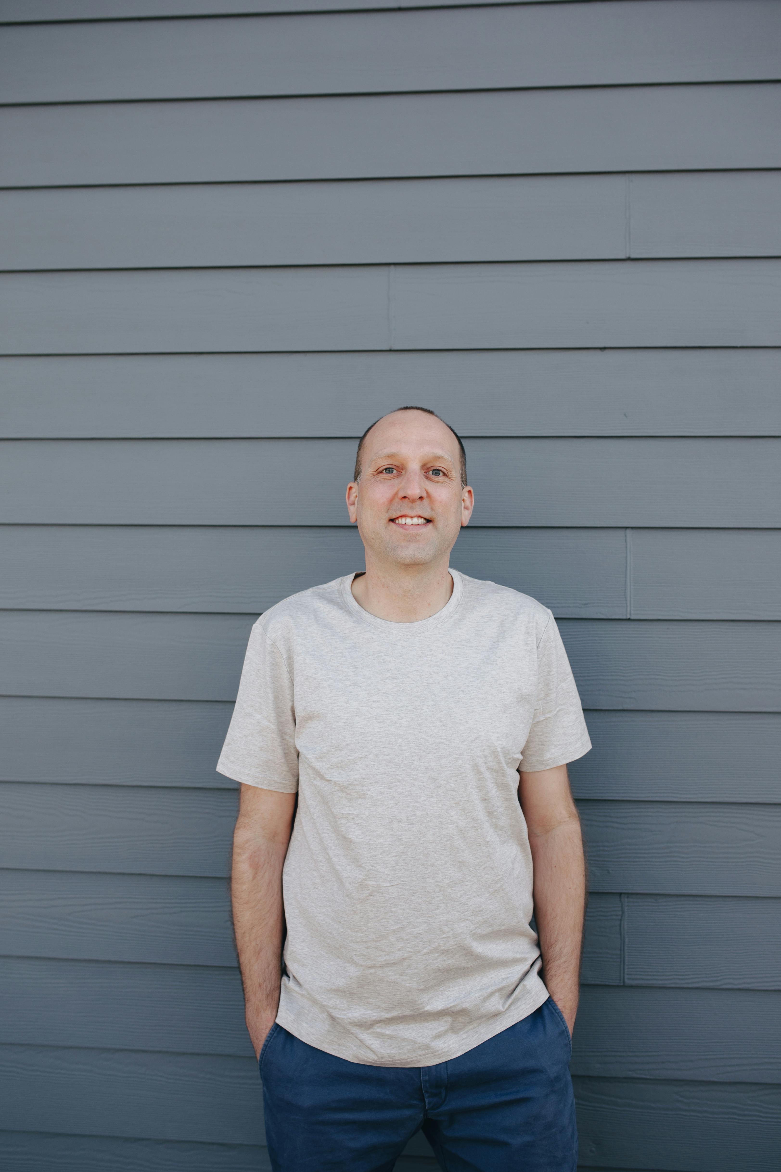 Casual portrait of a smiling man standing against a gray wall, outdoors during the day.