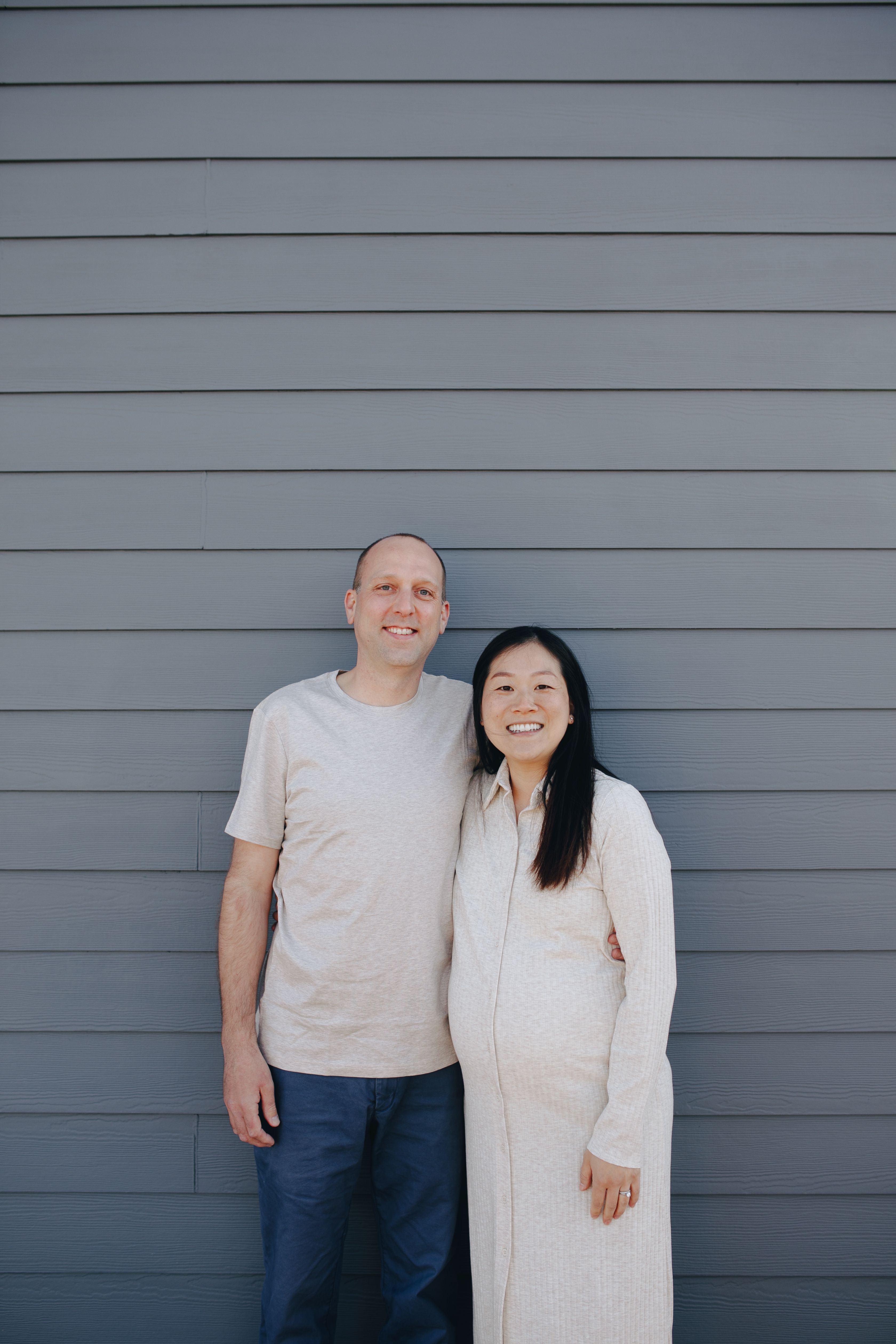 Smiling expectant couple posing together against a gray wall outdoors.
