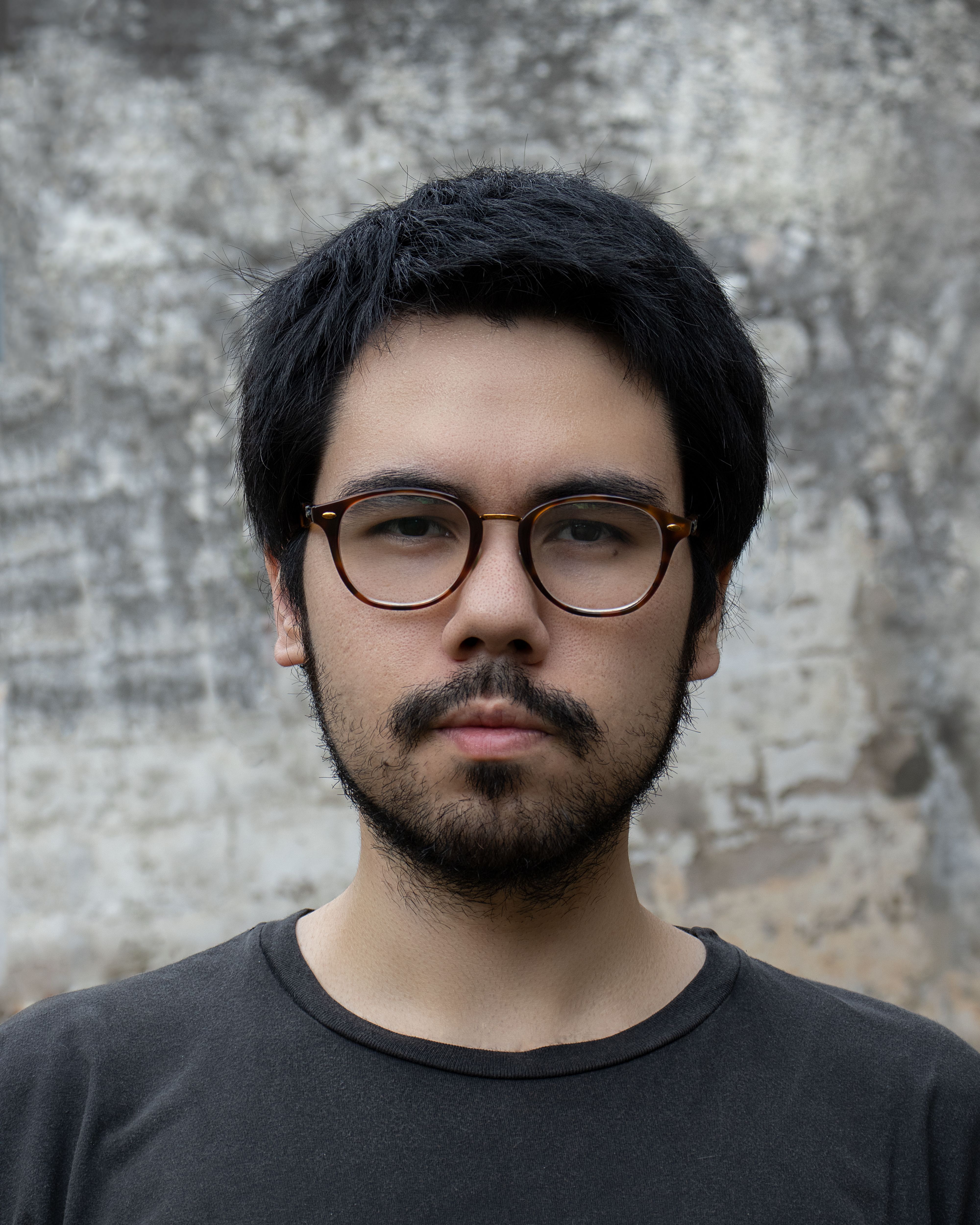Close-up portrait of an adult man with a beard and eyeglasses against a textured wall.