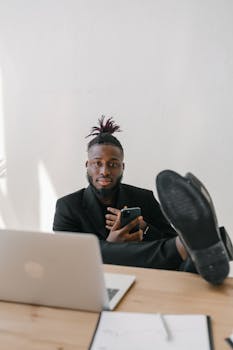 Young man in a suit relaxing at his desk with a laptop and phone in a modern office.